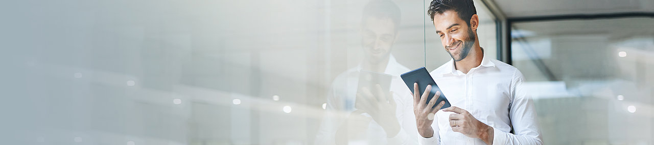 A man in business casual attire standing in an office window smiling at an iPad.
