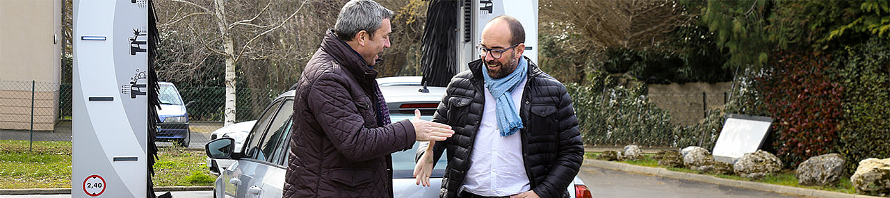 Two men dressed in fall attire shaking hands in agreement standing in front of a car using a car wash.