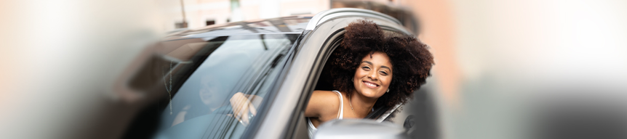 Woman smiling driving black crossover with her friend while sticking her head out of the window with wind blowing in her hair.