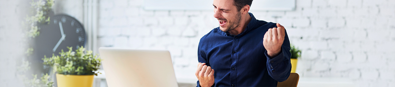 A man in a sunny room wearing a blue button down pumping his fist in joy while looking at laptop.